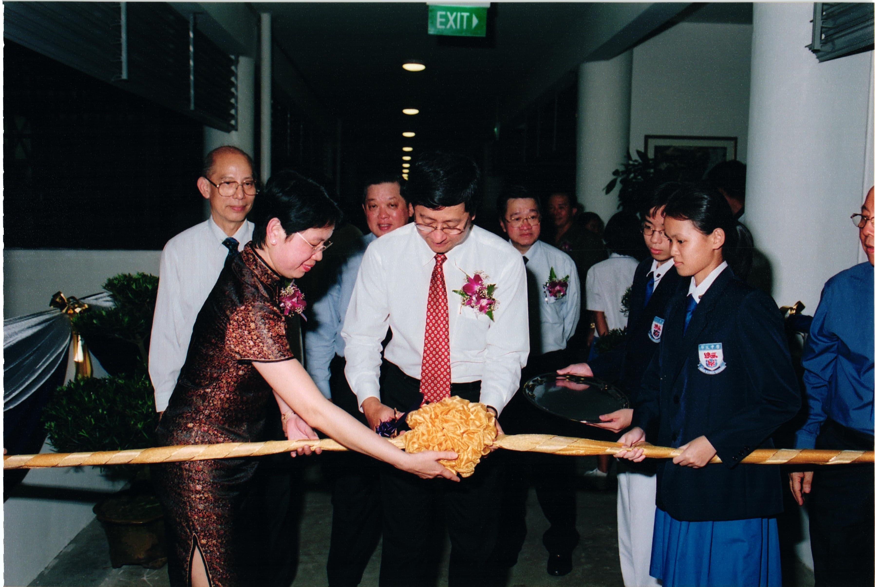 A man in a white shirt and red tie cuts a ribbon at a formal opening ceremony with several people watching.